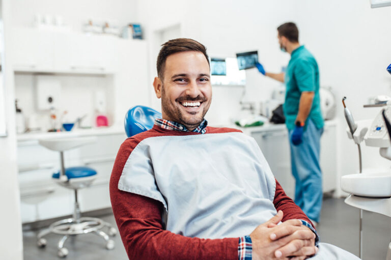 a male patient smiling in an operating chair with the assistant looking at an x-ray in the background