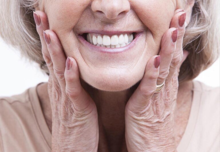Close-up of an older adult smiling with clean, well-aligned teeth, hands gently pressing against their cheeks. The person wears a light top and ring, with visible skin texture and painted nails.